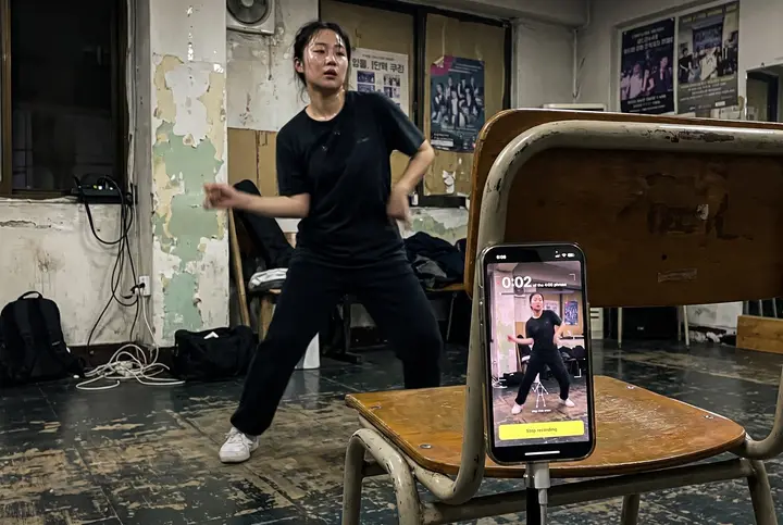 A dancer mid-move in a worn Seoul practice room at night, a propped phone running the app. AI-generated scene · real app screen